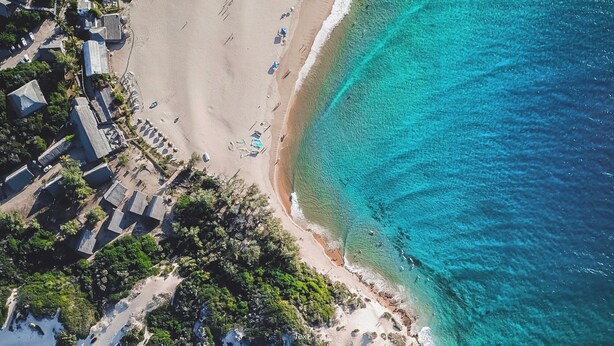 Vue aérienne d’une zone côtière avec une plage, des eaux turquoise et limpides, et un petit groupe de bâtiments adossés à un promontoire rocheux.