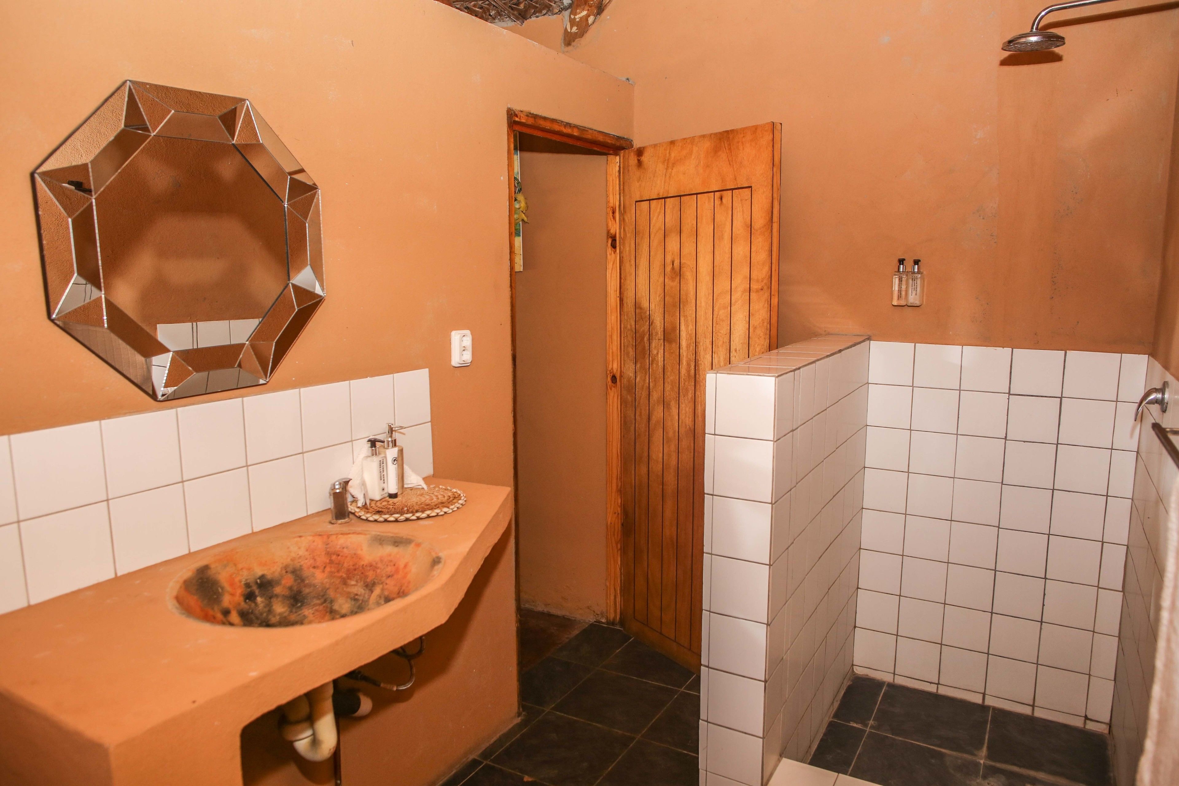 Un baño con puerta de madera, un lavabo con artículos de aseo y una zona de ducha con azulejos blancos.