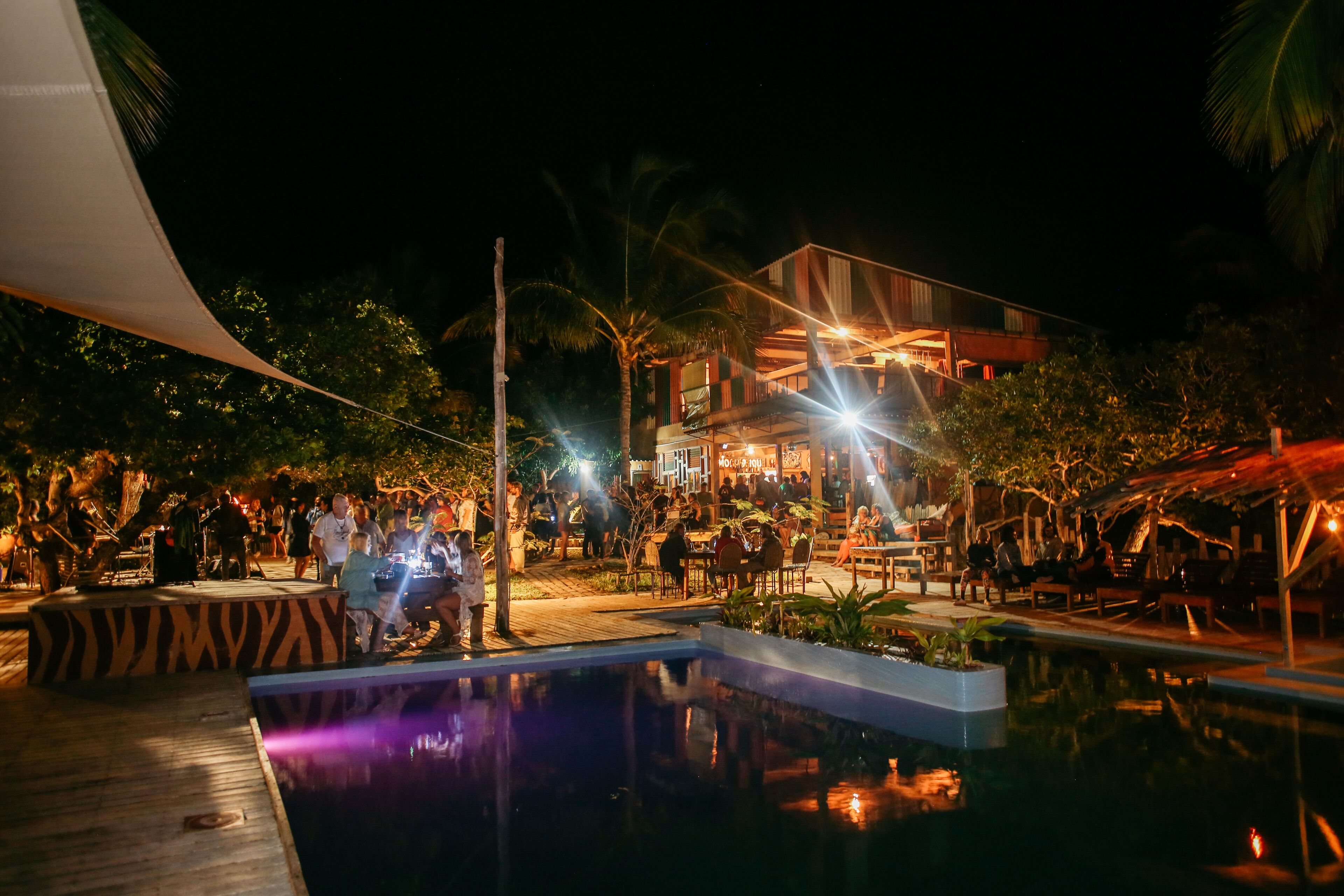 Área de piscina externa à noite com um deck de madeira, cercada por palmeiras e um edifício com janelas iluminadas.