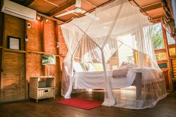 Una habitación con una cama bajo una mosquitera, paredes de madera, una mesita con una lámpara y un tapete rojo.