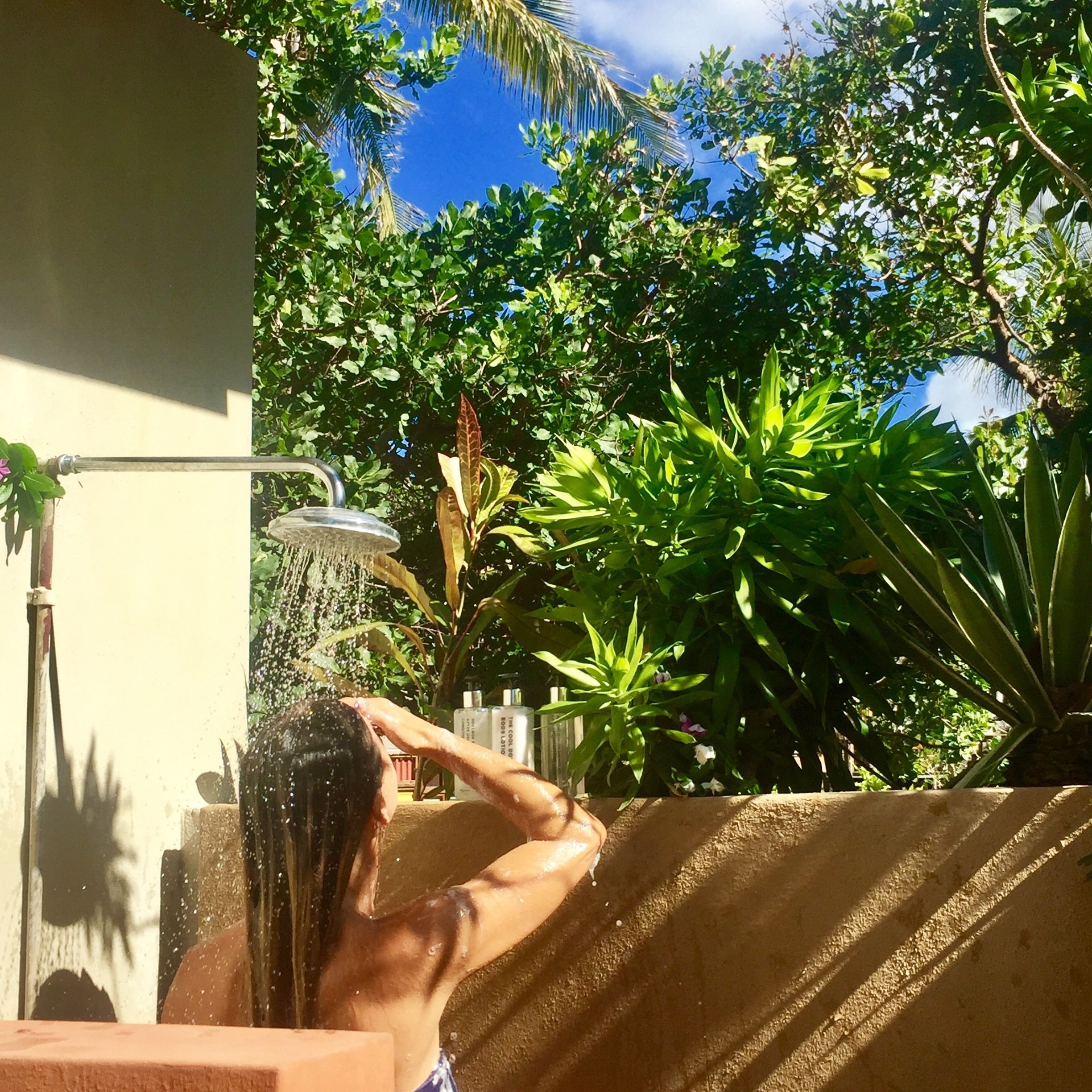 A person taking a shower in a covered outdoor area with a view of lush green trees and a clear blue sky.