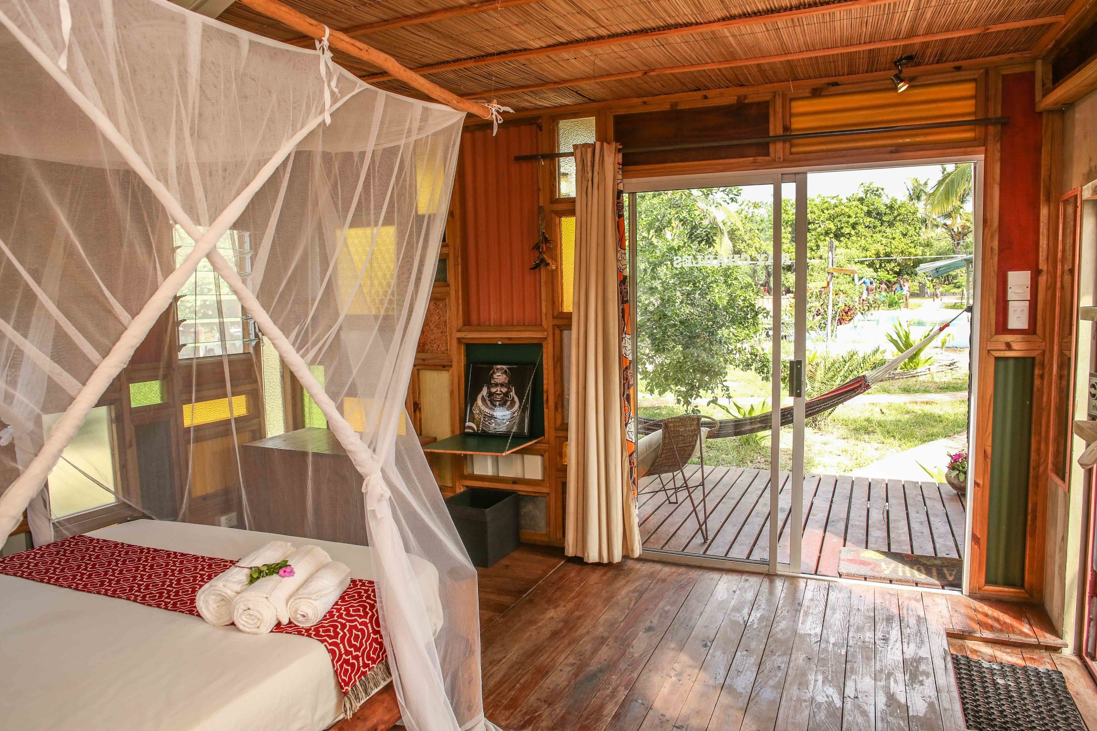 A room with a bed, a mosquito net, a small table with a statue, and a view of the outdoors through a glass door.