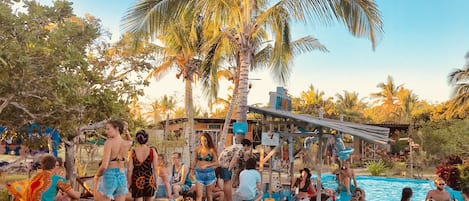 A poolside area with people relaxing and swimming, surrounded by palm trees and a wooden deck.