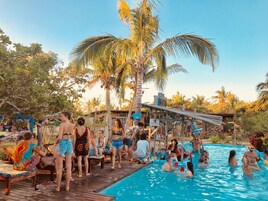 Zona junto a la piscina con gente relajándose y nadando, rodeada de palmeras y una terraza de madera.