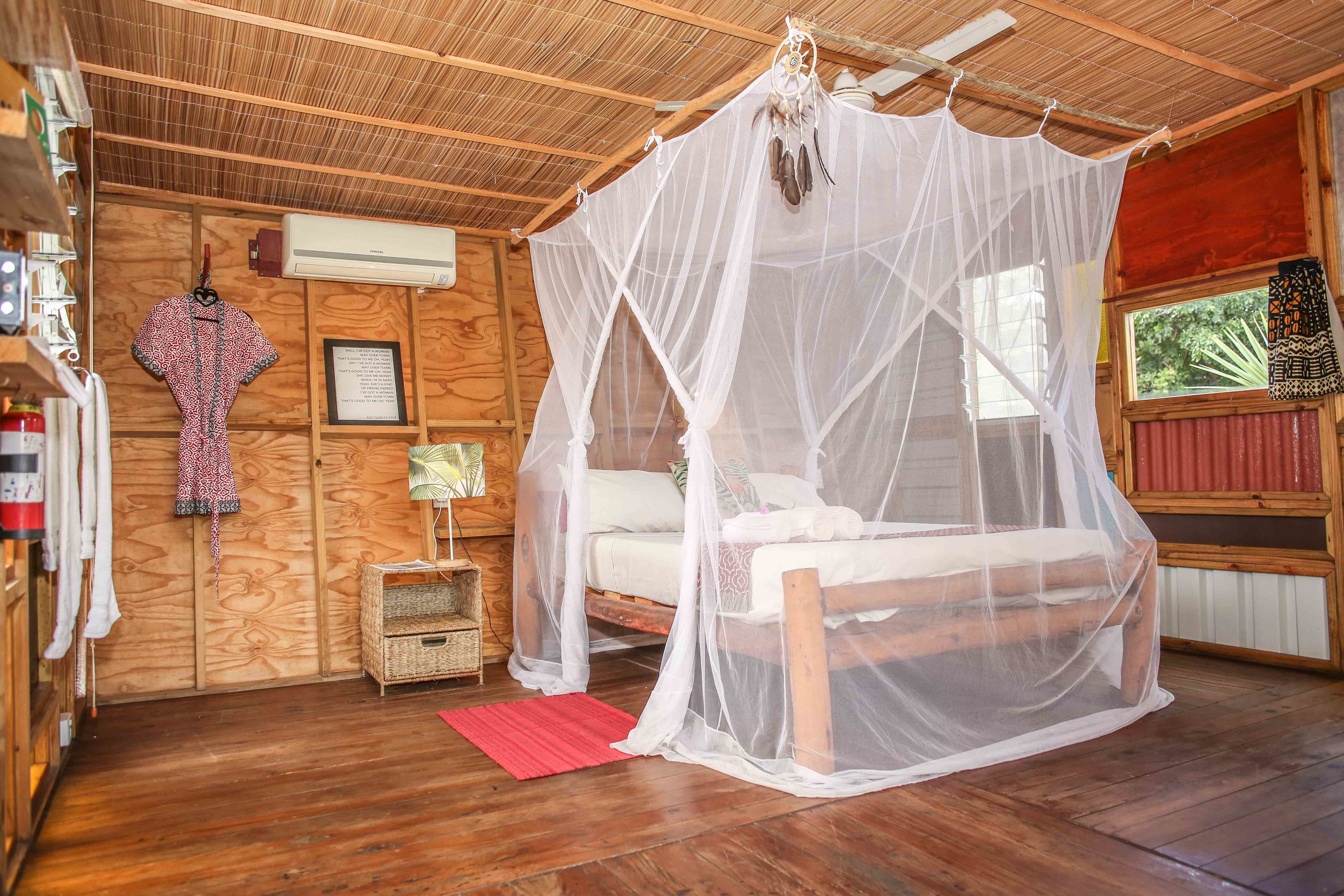 A wooden cabin room with a bed under a mosquito net, a small bedside table, and a window with a view of greenery.