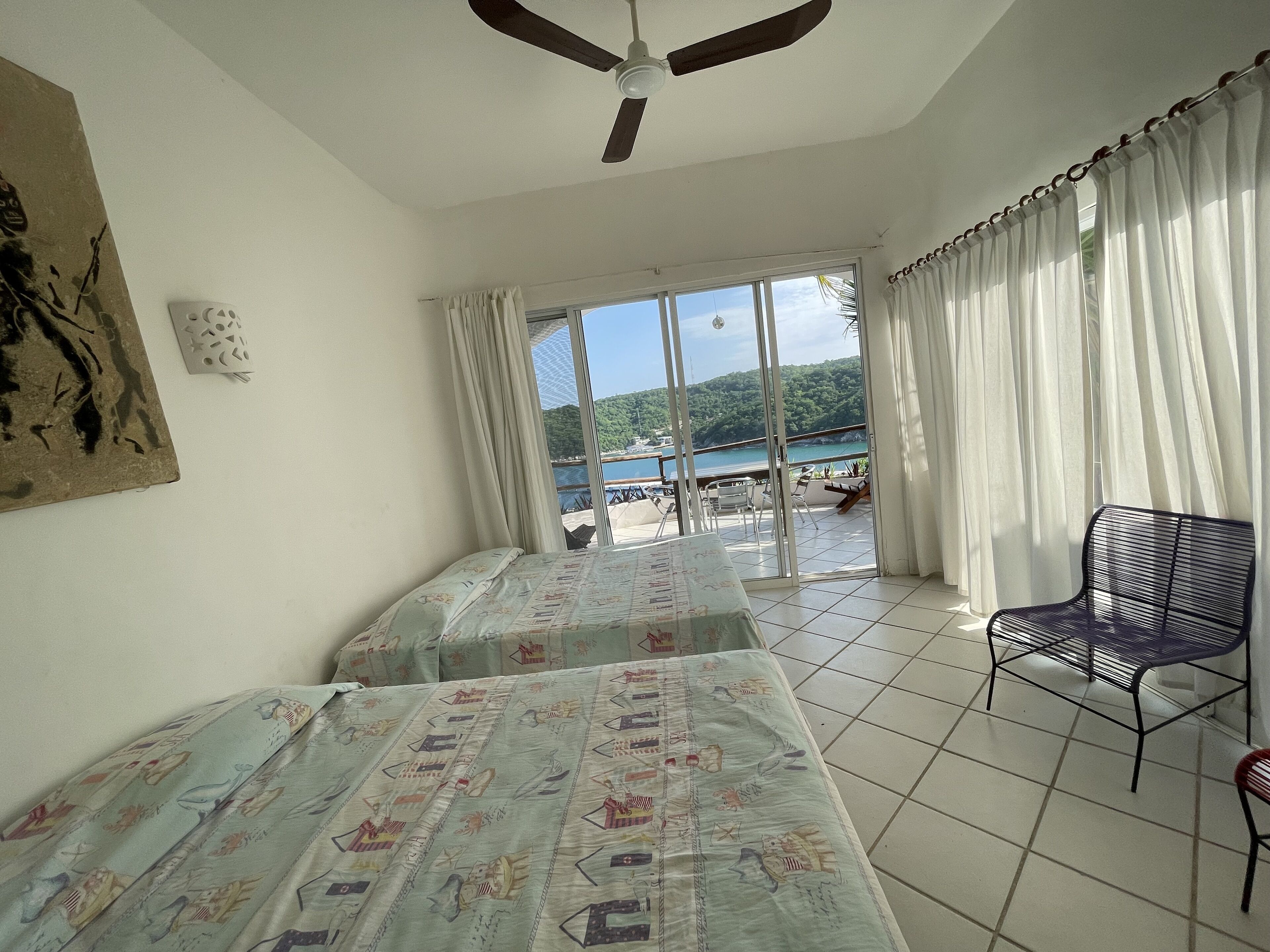 A room with two beds, a ceiling fan, a chair, and a view of a beach and mountains through large windows.