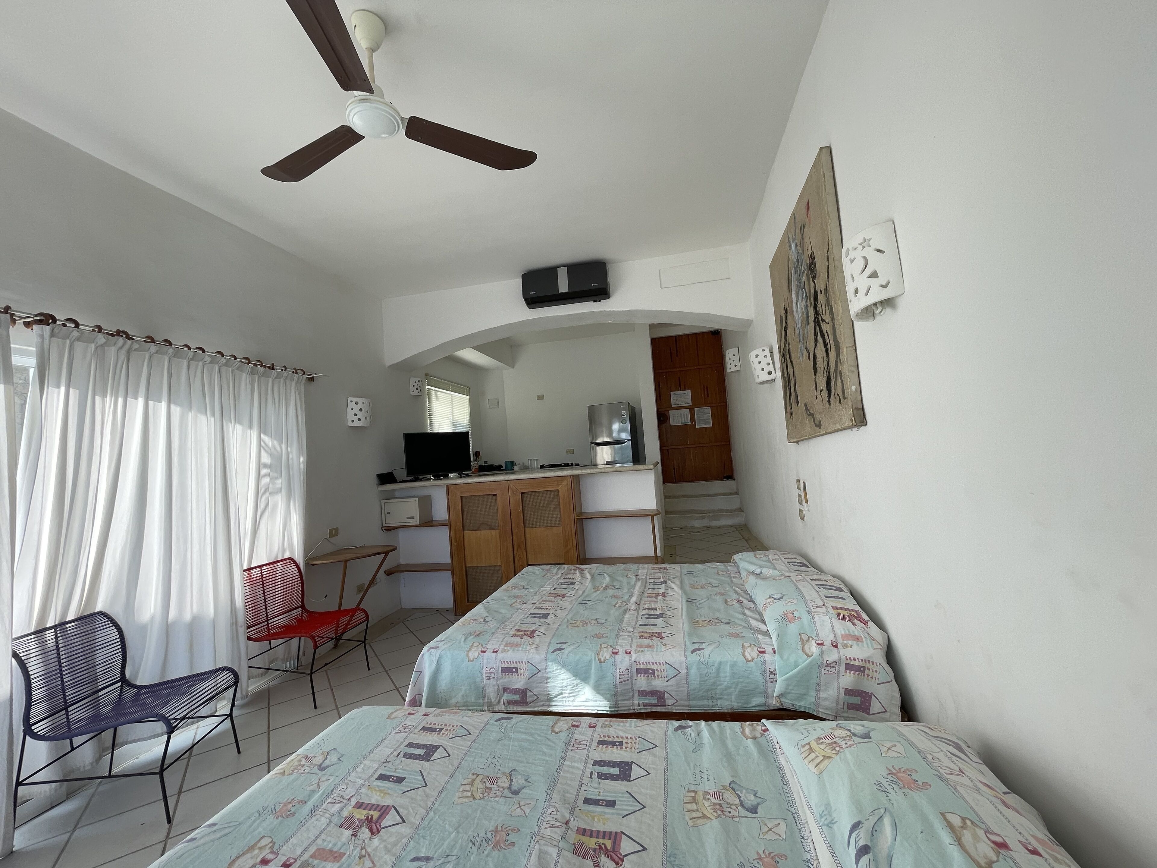 Un dormitorio con dos camas, un escritorio, una silla, un ventilador de techo y una zona de cocina al fondo.