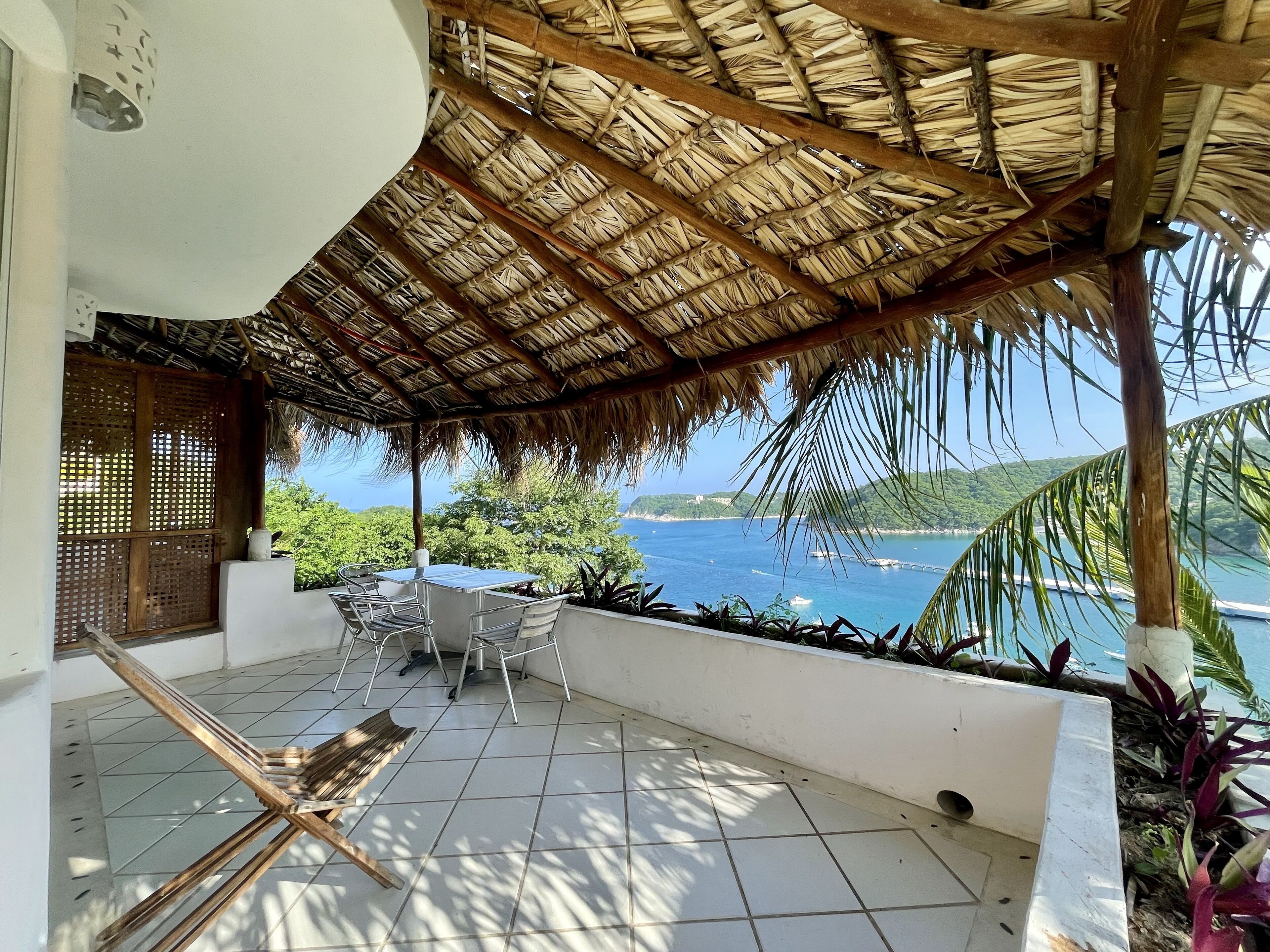 A covered patio with a thatched roof, a dining table with chairs, and a view of the sea and hills.