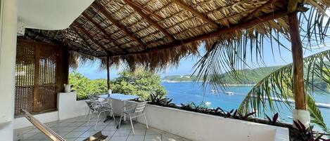 A covered patio with a thatched roof, a dining table with chairs, and a view of the sea and hills.