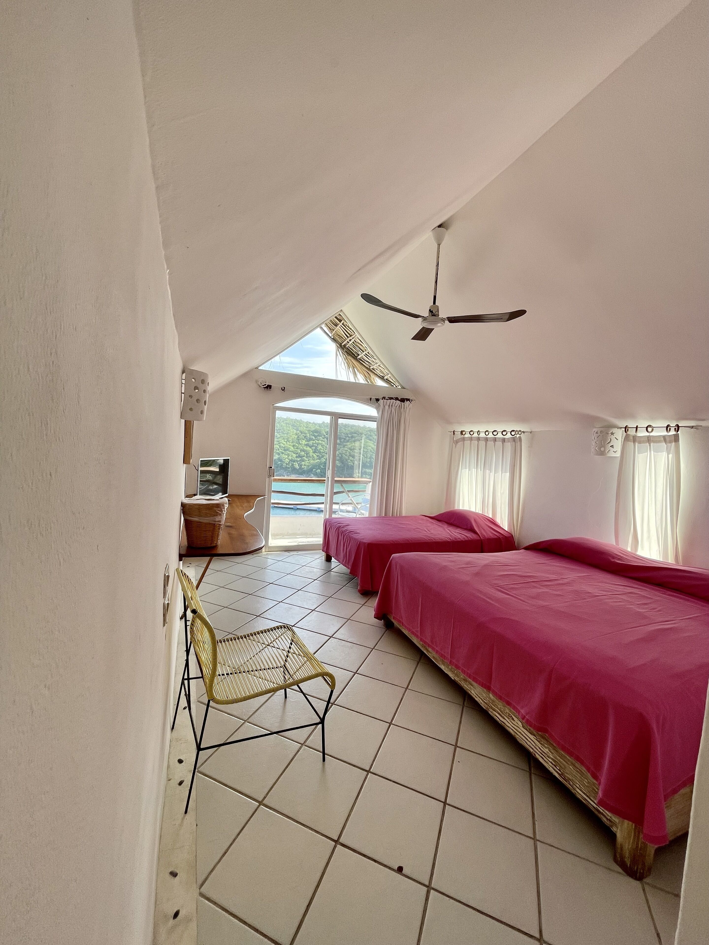 Una habitación con dos camas, ventilador de techo, ventana con cortinas, una silla y vista a una piscina.