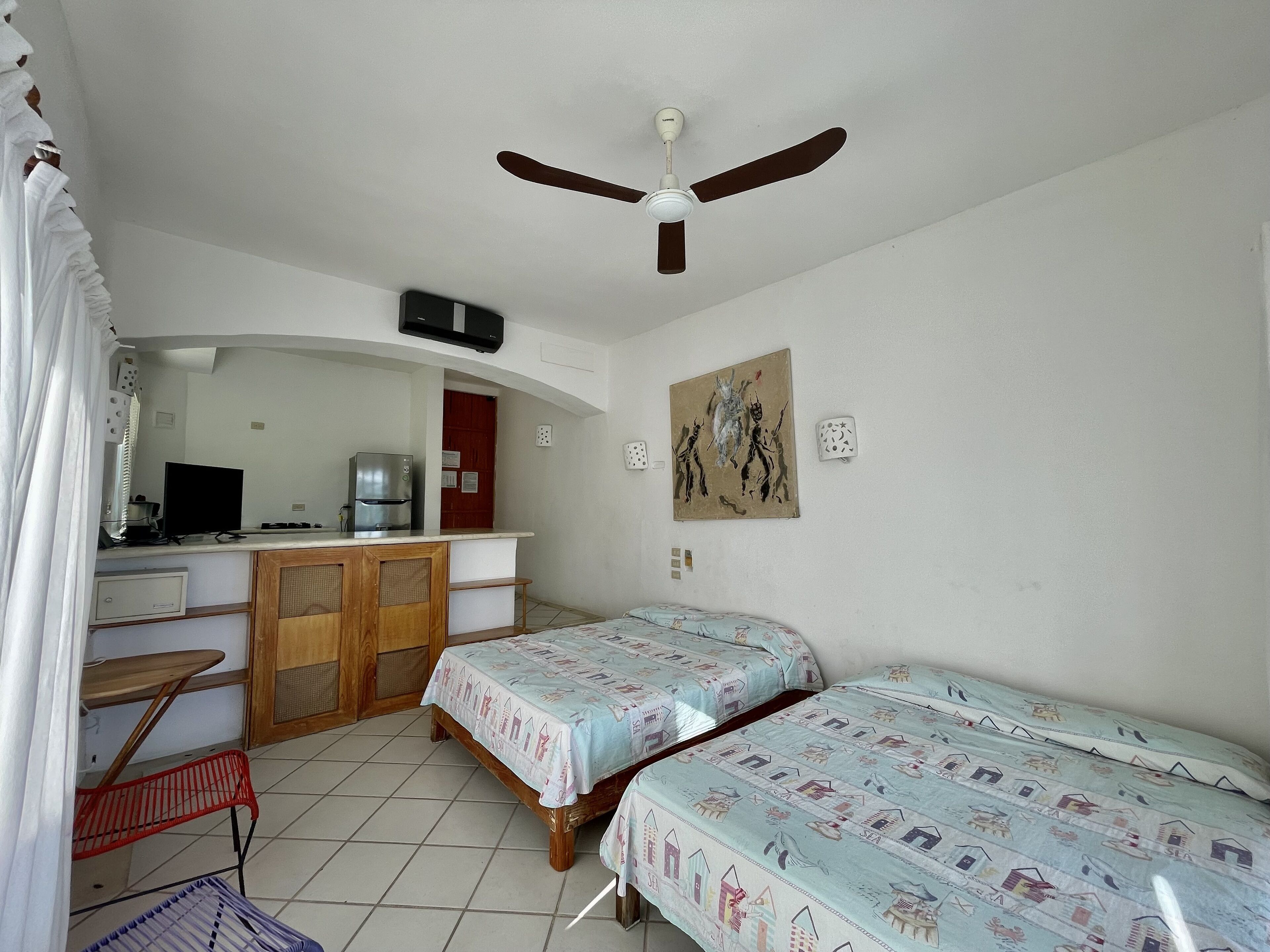 A bedroom with two beds, a desk, a chair, a wardrobe, a ceiling fan, and a painting on the wall.