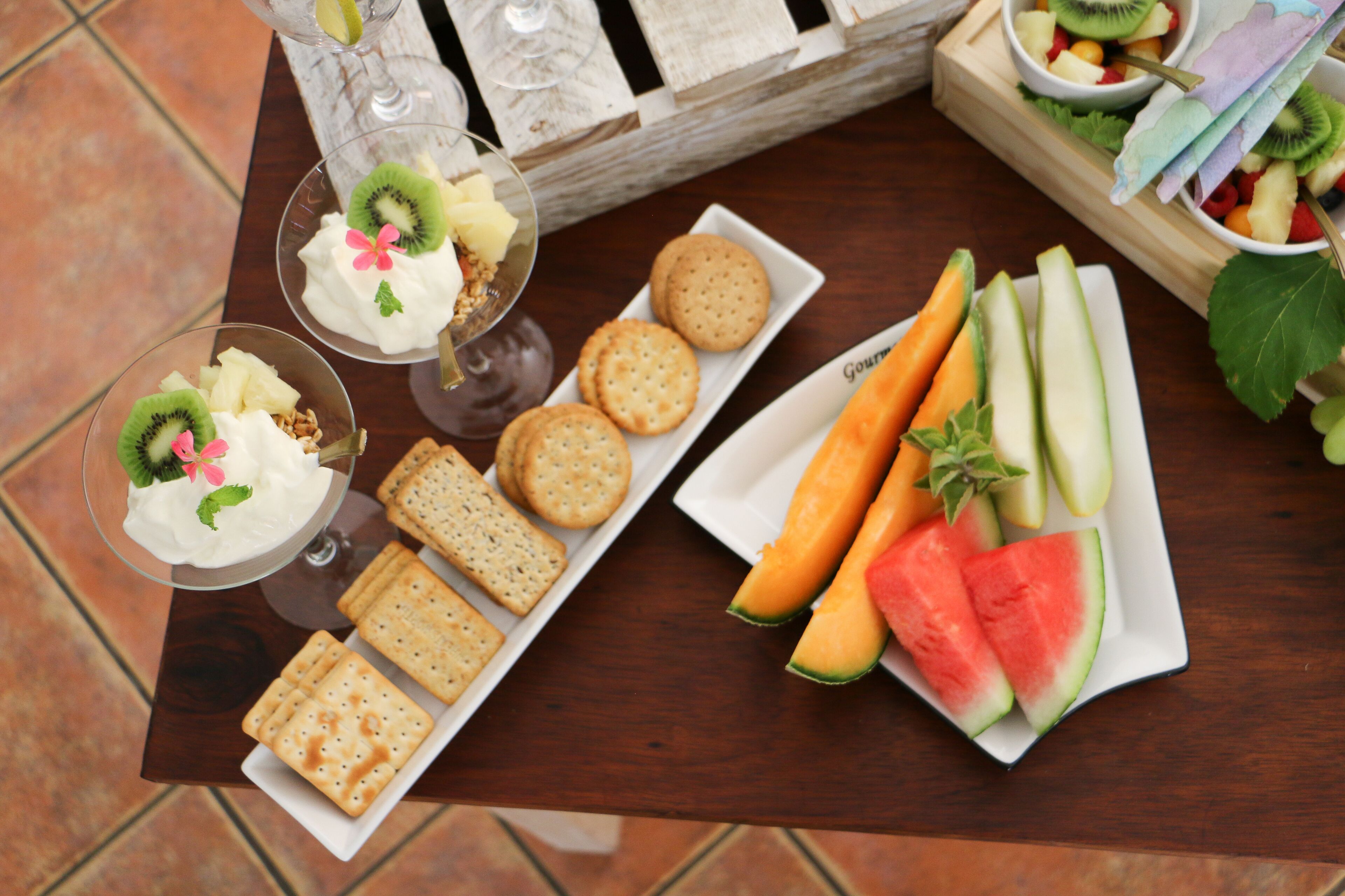 A table with a variety of snacks including crackers, sliced fruits, and a dessert with whipped cream and kiwi.