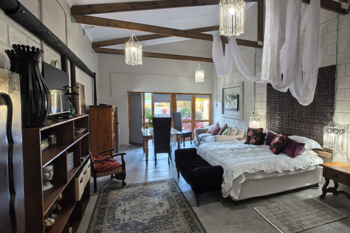 A bedroom with a large bed, a wooden nightstand, a bookshelf, and a desk. There is a window with a view of the outside.