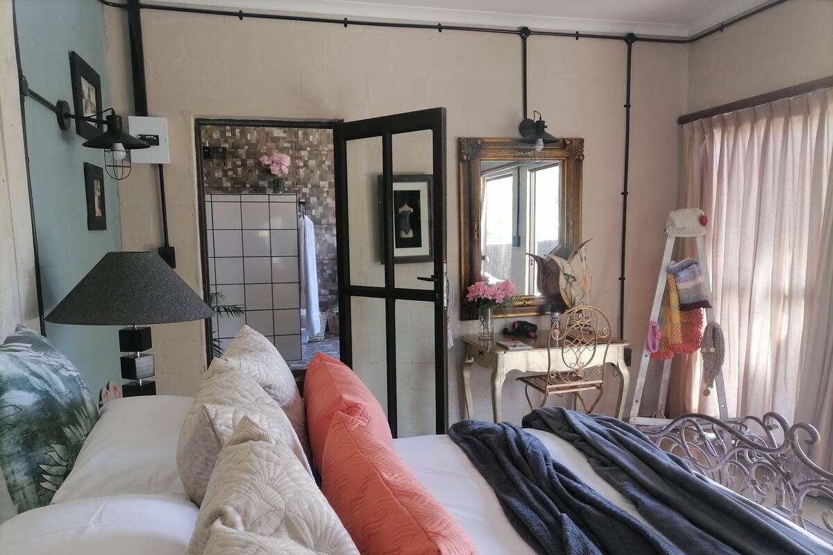 A neatly made bed with pillows, a bedside lamp, a mirror, and a window with curtains.