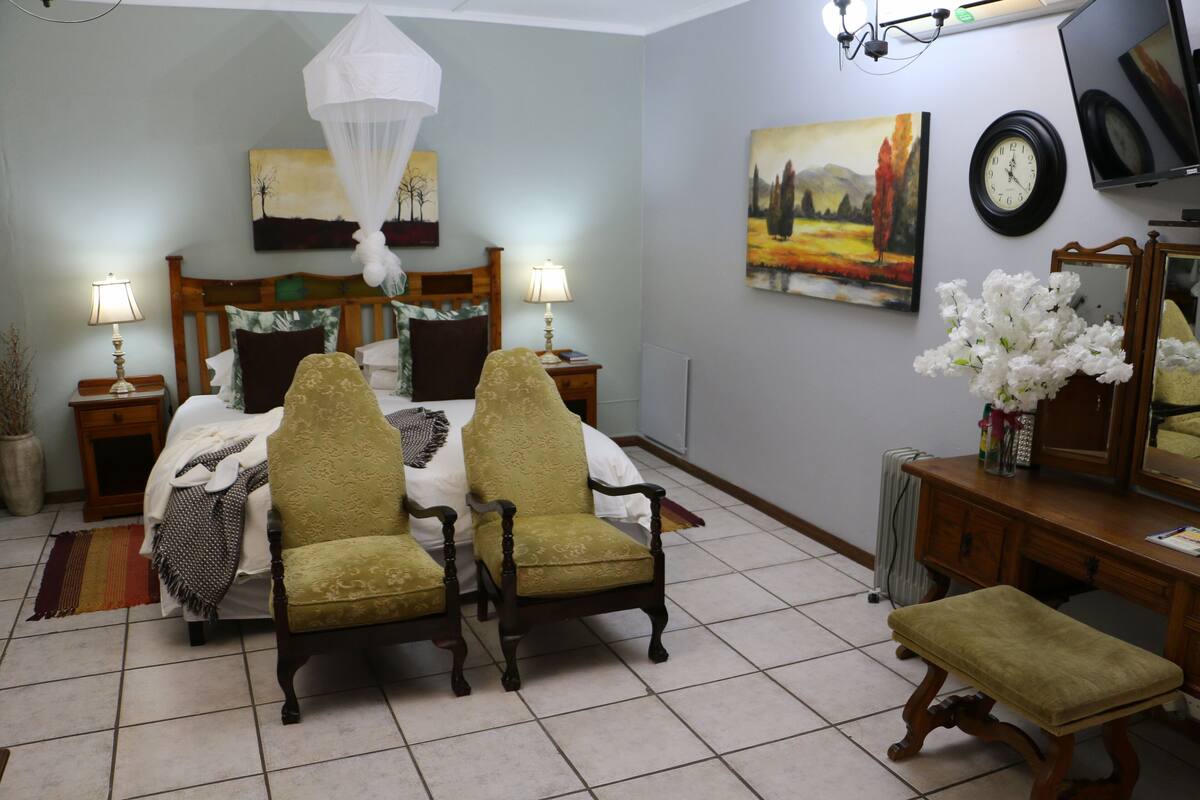 A bedroom with a bed, two chairs, a bench, a bedside table with lamps, a bedside table with a clock, a dresser, a mirror, and wall art.