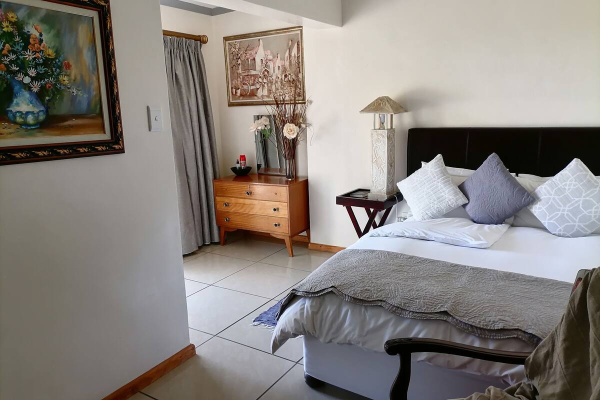 A bedroom with a bed, a nightstand, a dresser, a painting, and a window with curtains.