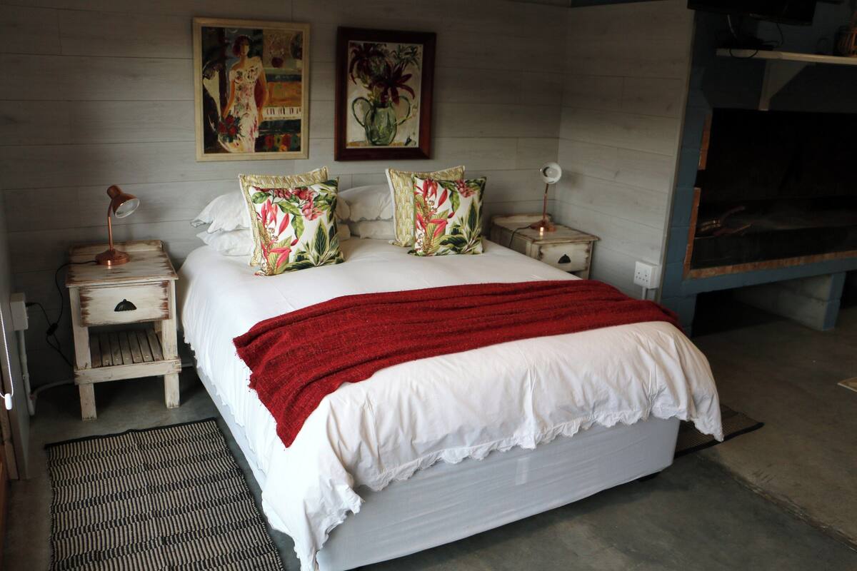 A bedroom with a large bed, two nightstands, a red throw blanket, floral pillows, and two wall-mounted paintings.