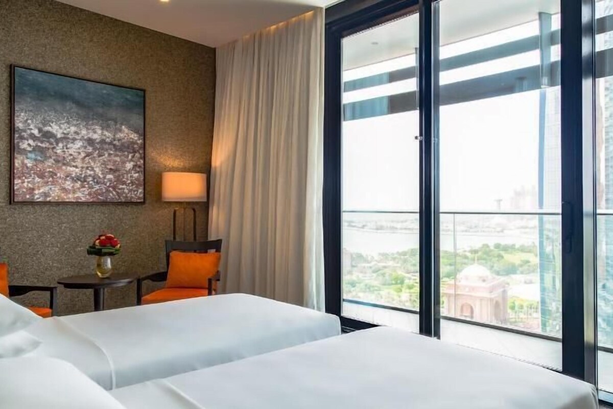 A hotel room with two beds, a cityscape painting, a lamp, and a view of buildings through large windows.