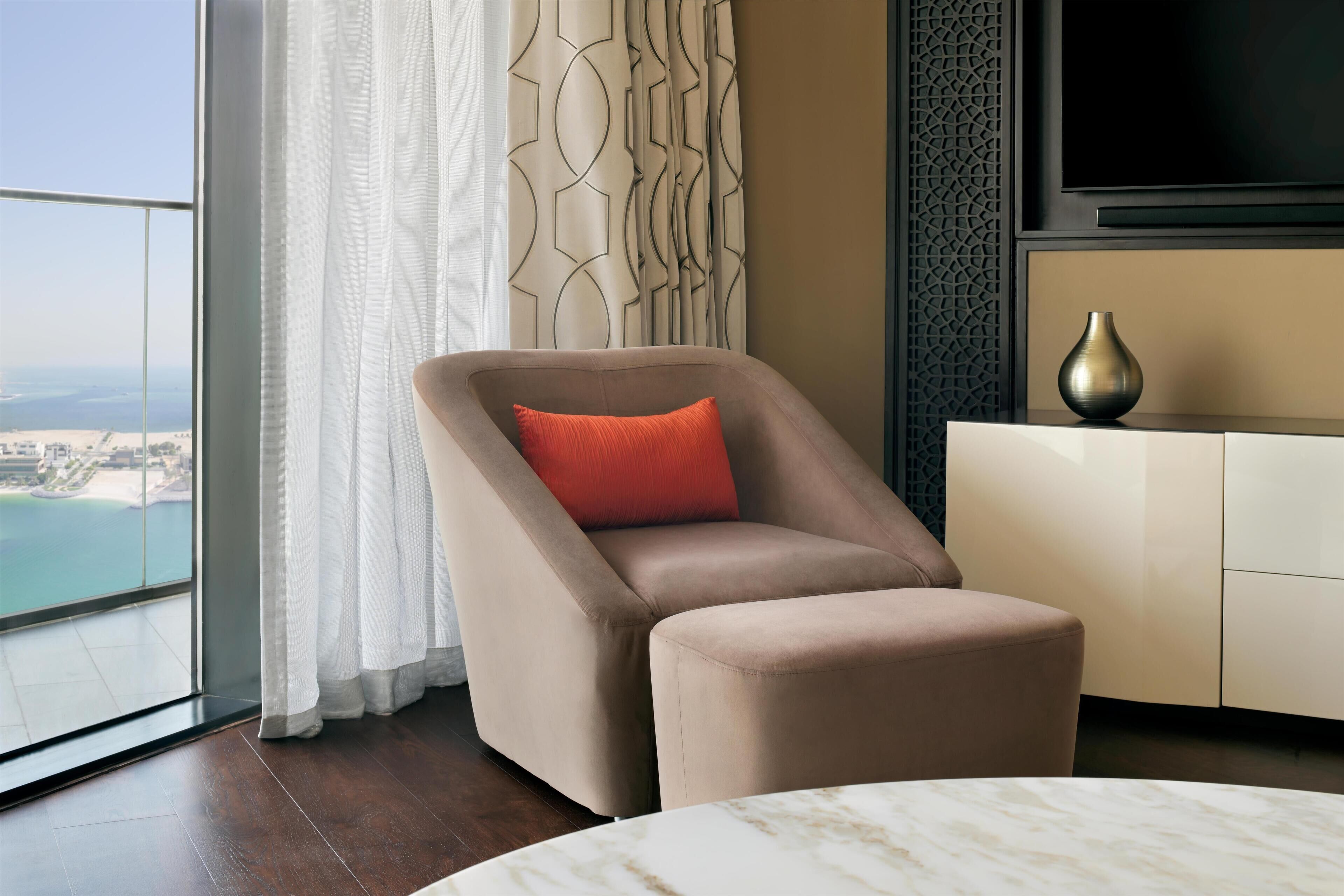 Habitació d'hotel moderna amb un gran finestral que ofereix vistes a la platja, un sofà còmode amb coixí vermell, una taula de cafè de marbre i un moble de televisió de disseny minimalista.