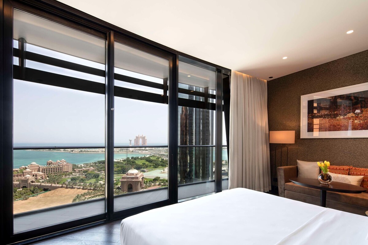 A hotel room with a large window offering a view of a coastal cityscape, a bed, a nightstand, and a sofa.