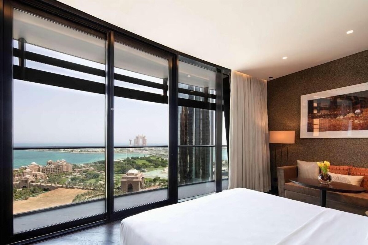 A hotel room with a large window, a bed, a sofa, and a view of a cityscape and body of water.