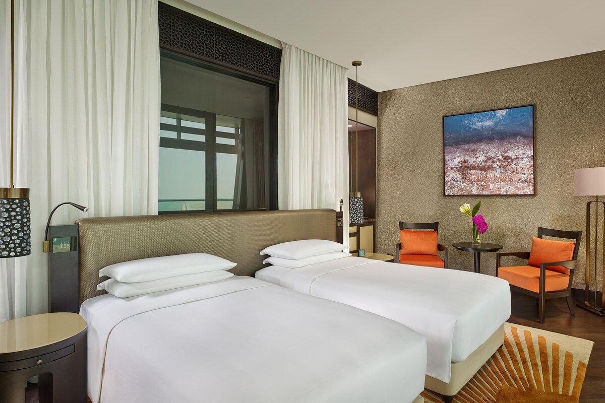 A hotel room with two beds, a large window with curtains, a painting on the wall, and a small table with a lamp.