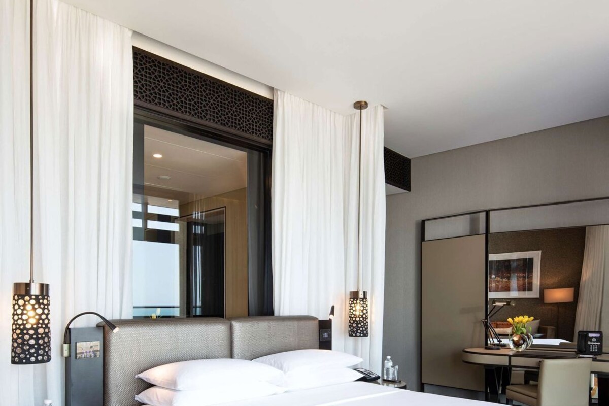 A modern hotel room with a large bed, a desk, a chair, a mirror, and a view of the city.