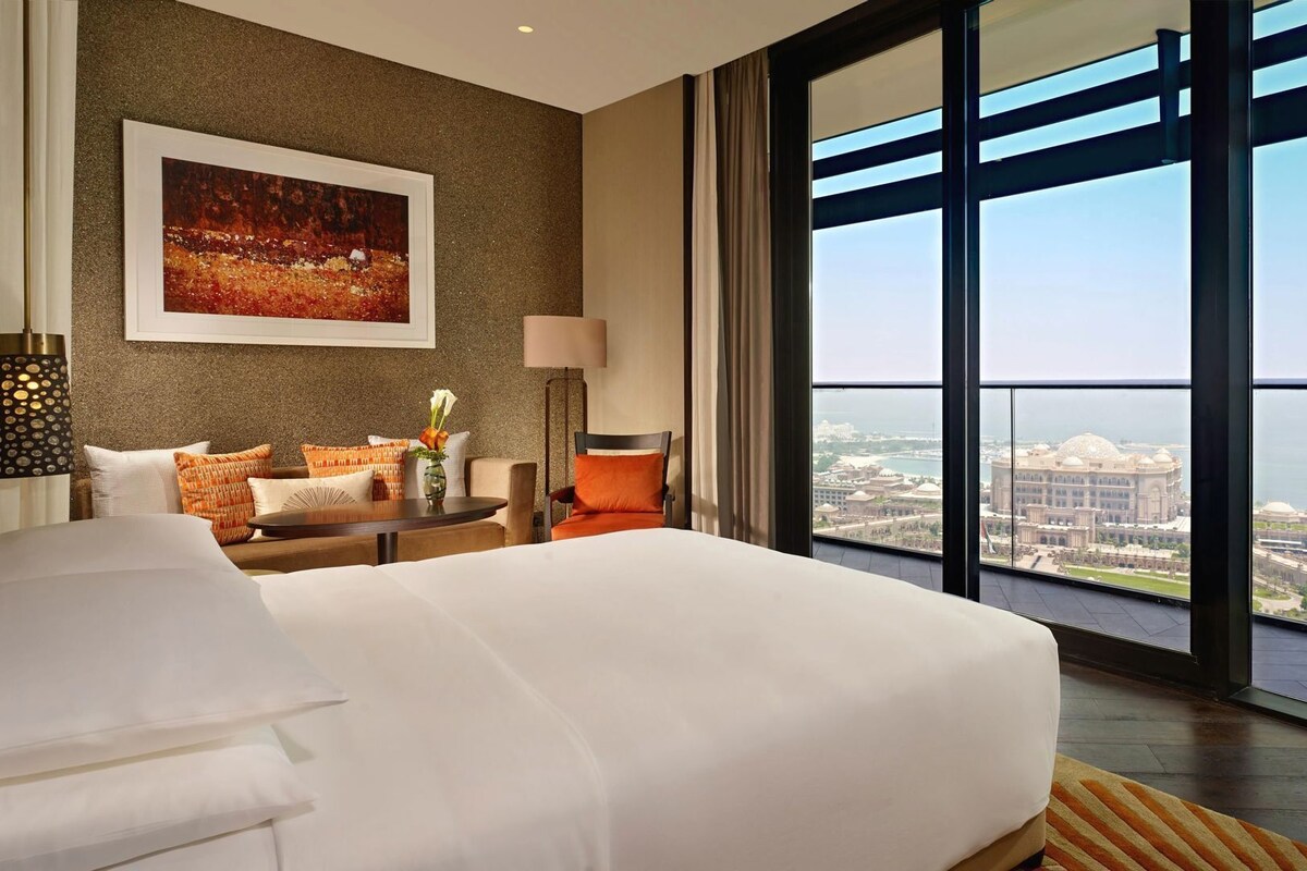 A hotel room with a large bed, a desk, a chair, a lamp, a painting, and a large window with a city view.