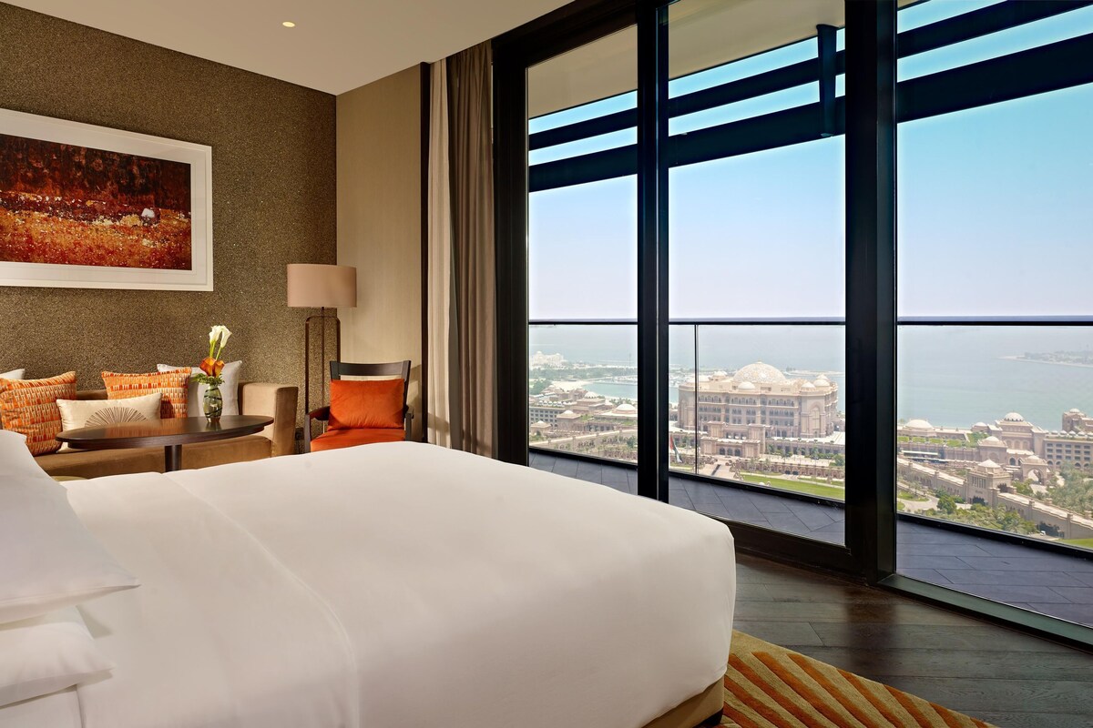 A hotel room with a large bed, a sofa, a table with a vase of flowers, a painting on the wall, and a view of a cityscape through large windows.