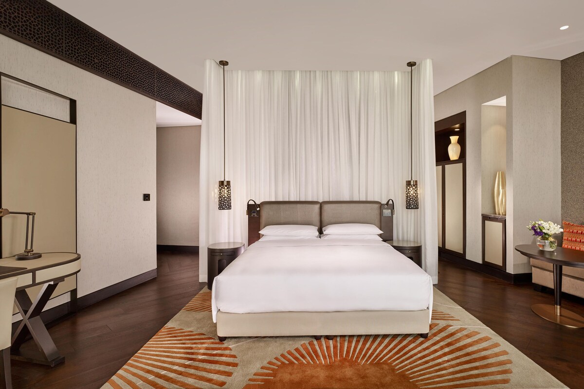 A modern hotel room with a large bed, a desk, a chair, and a small table with a vase.