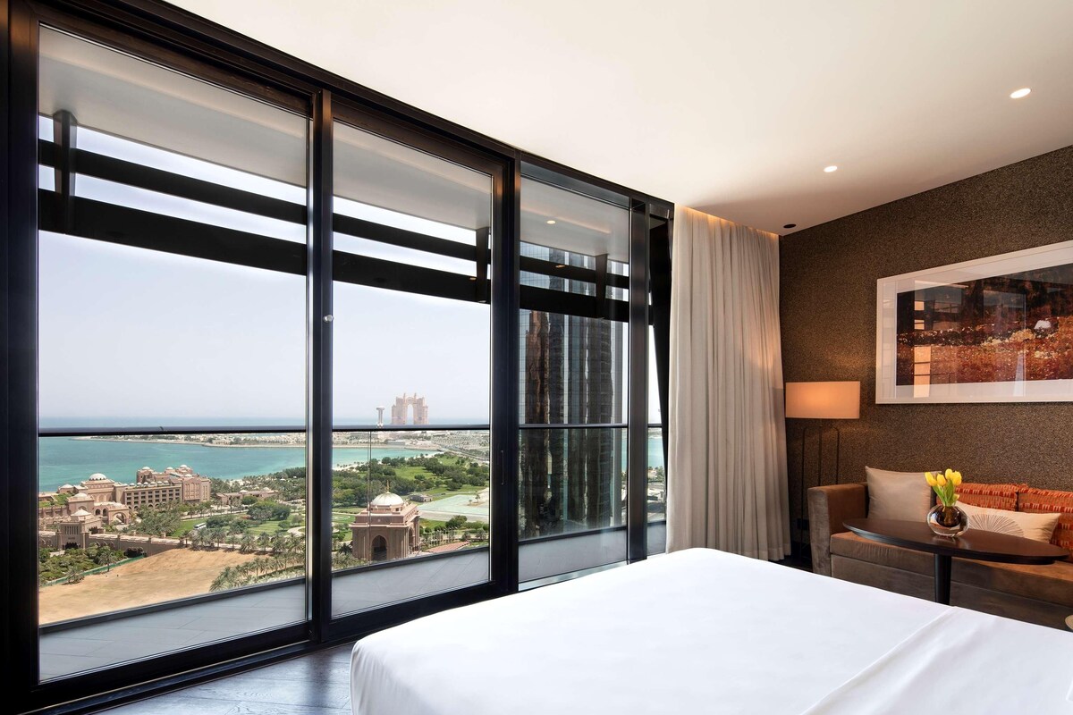 A hotel room with a large window offering a view of a coastal cityscape, a bed, a nightstand, and a sofa.