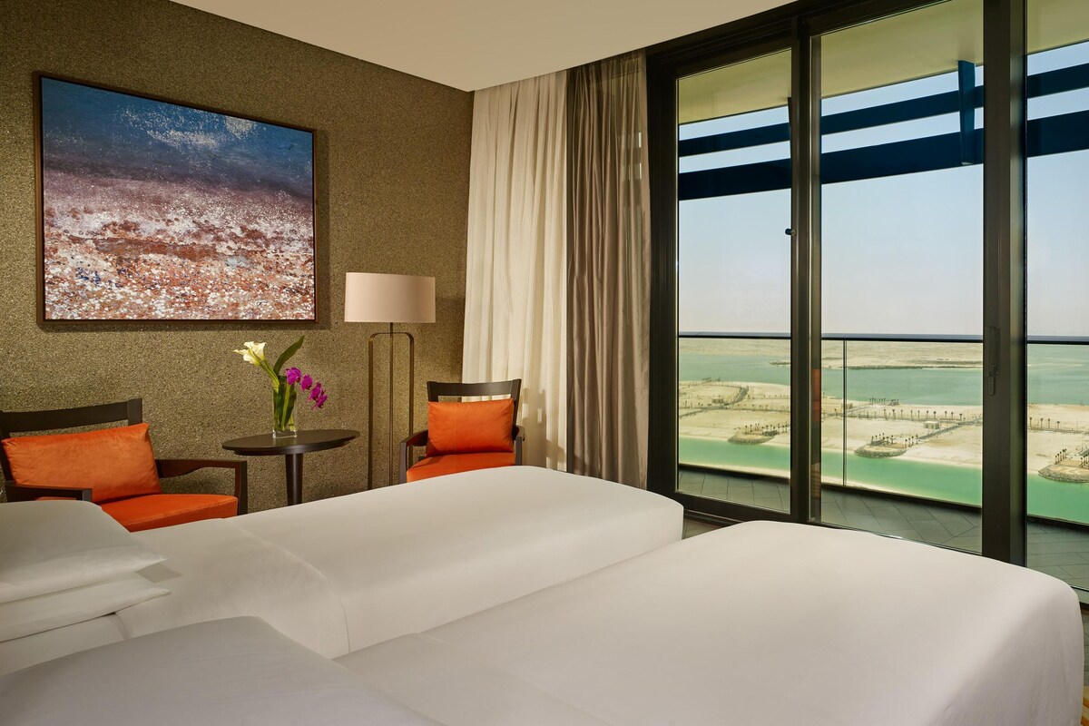 A hotel room with a large bed, two chairs, a table with a vase of flowers, a painting on the wall, and a balcony with a view of the beach and water.