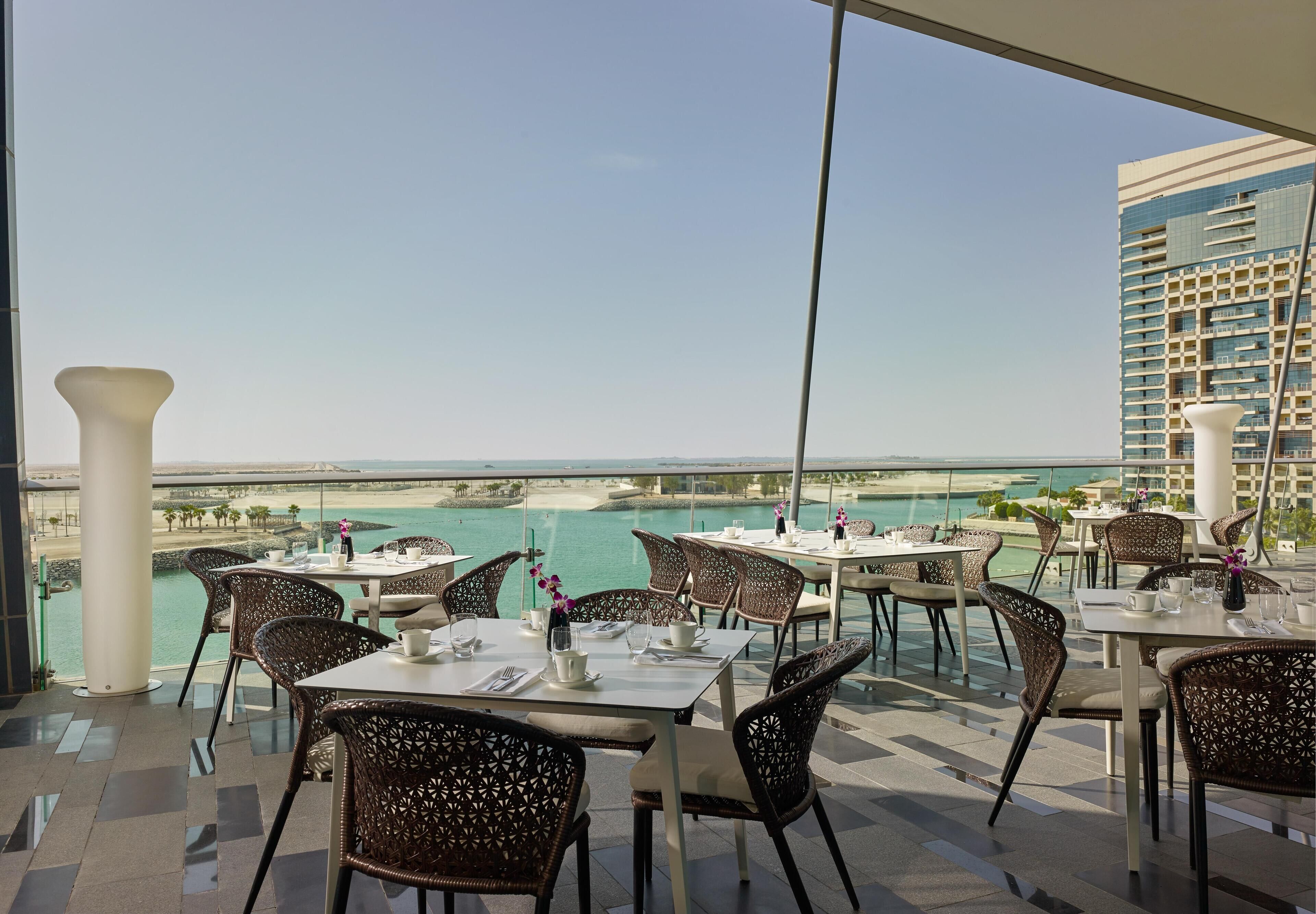 A restaurant with tables and chairs arranged on a terrace overlooking a beach and the sea.
