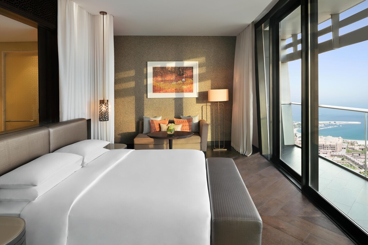 A hotel room with a large bed, a seating area with a table, and a view of the city and sea.