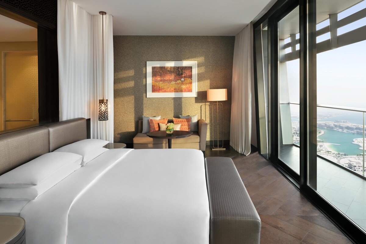 A modern hotel room with a large bed, a seating area with a table, and a view of a body of water through large windows.