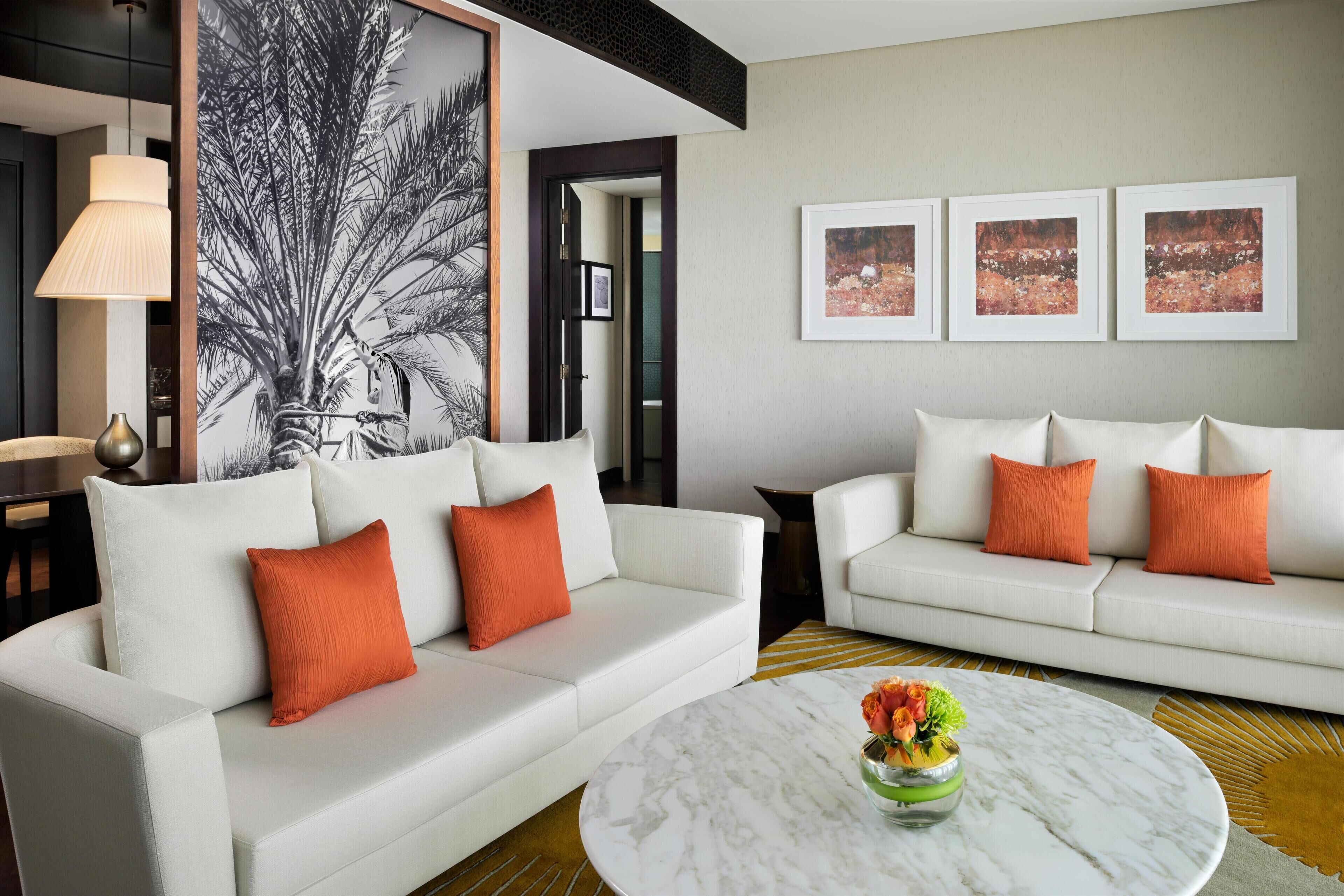 A modern living room with two white sofas, one with orange cushions, a marble coffee table with flowers, and a large abstract painting on the wall.