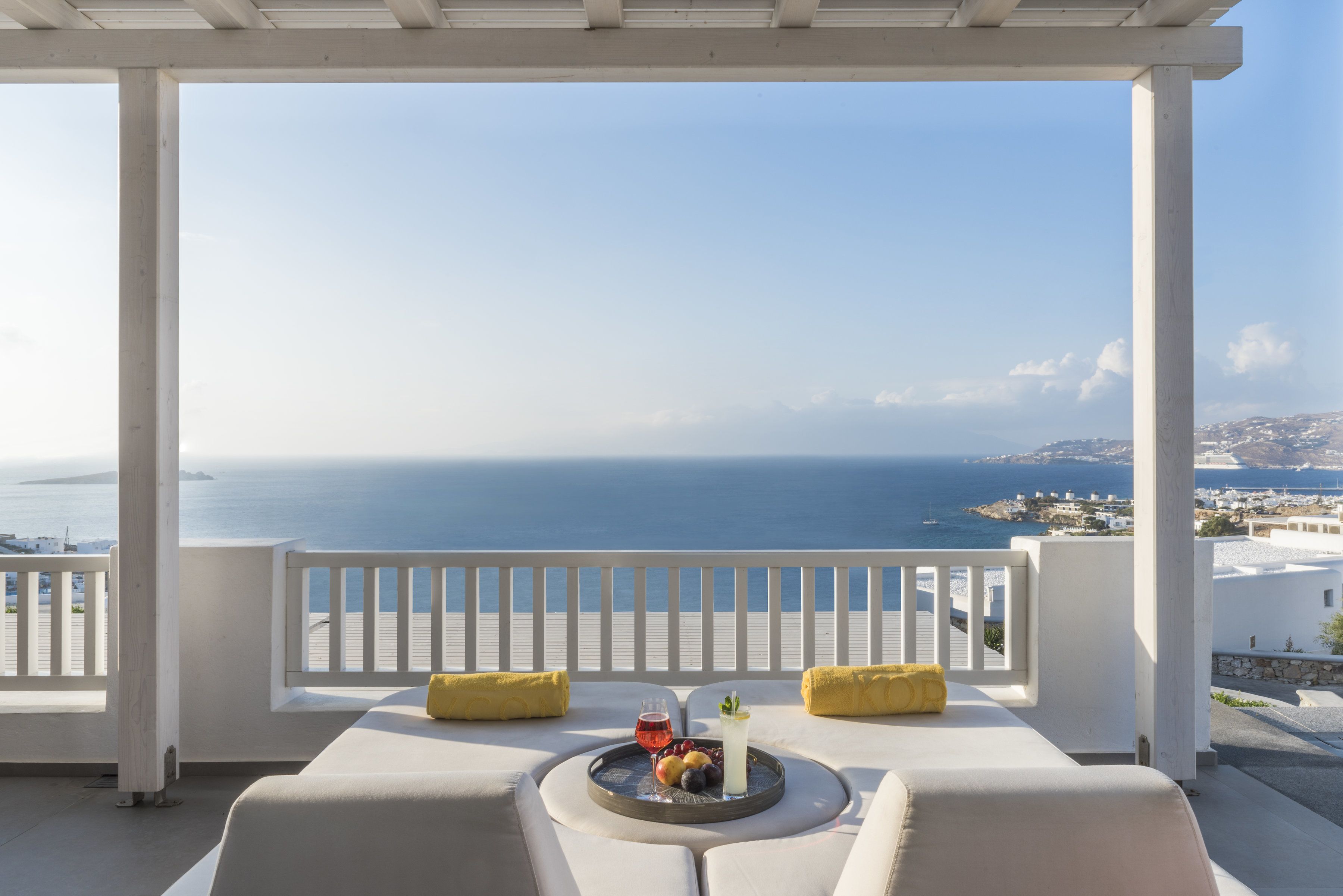 Un balcon avec vue sur la mer, un plateau avec des fruits et une boisson, et des meubles blancs agrémentés de coussins jaunes.