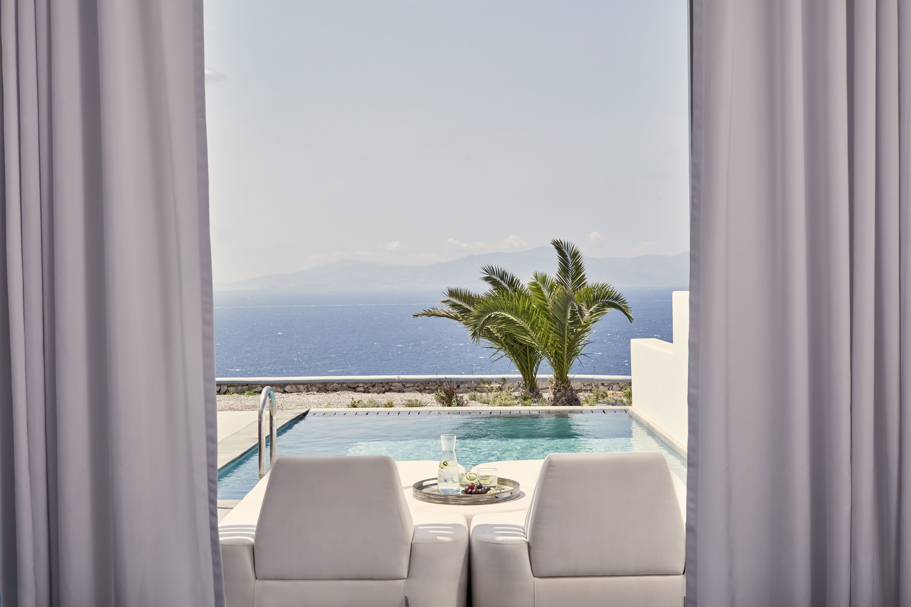 Vista de la piscina con mesa y sillas, una palmera y vistas al mar y las montañas.