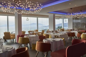 A restaurant with round tables, red and yellow chairs, and a view of the sea.