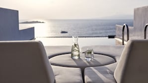 A round table with a vase and two glasses, overlooking a body of water and distant land. - Myconian Korali Relais & Chateaux (Mykonos)