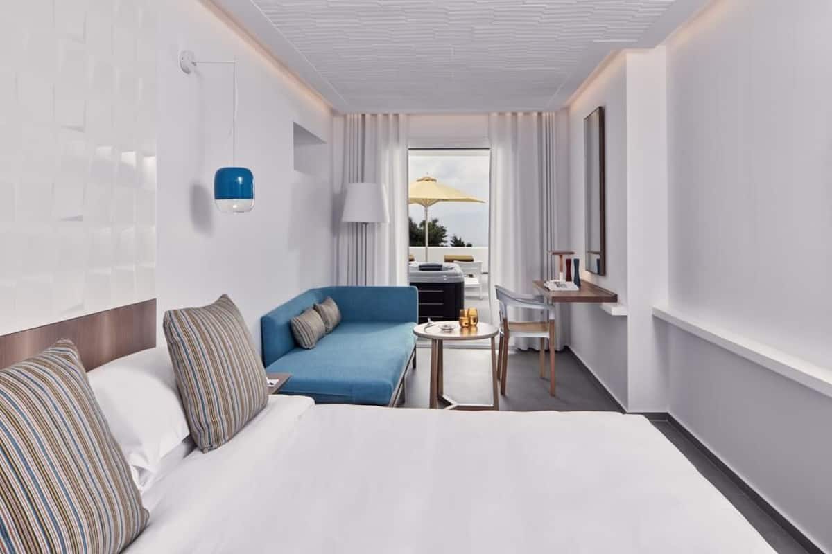 Ein Hotelzimmer mit einem Bett, einem blauen Sofa, einem kleinen Tisch, einem Schreibtisch, einer Lampe und einem Fenster mit Vorhängen.