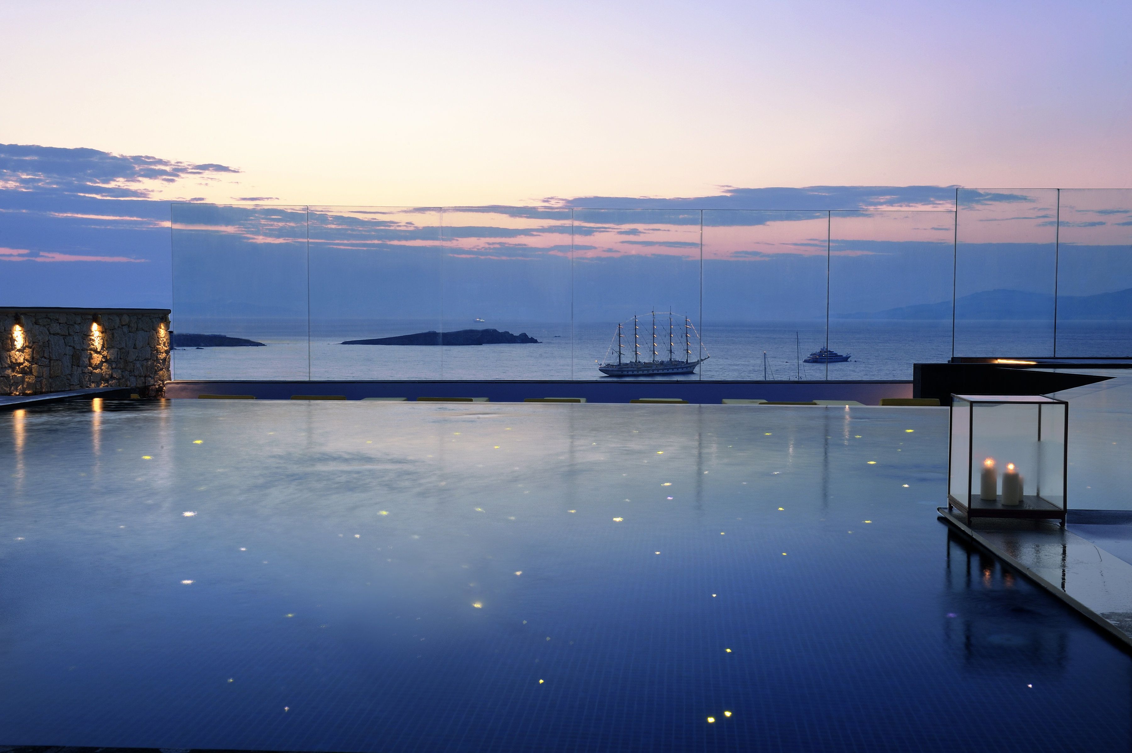 A serene infinity pool with a clear view of the sea and boats, a stone wall with lights, and a glass-enclosed seating area.