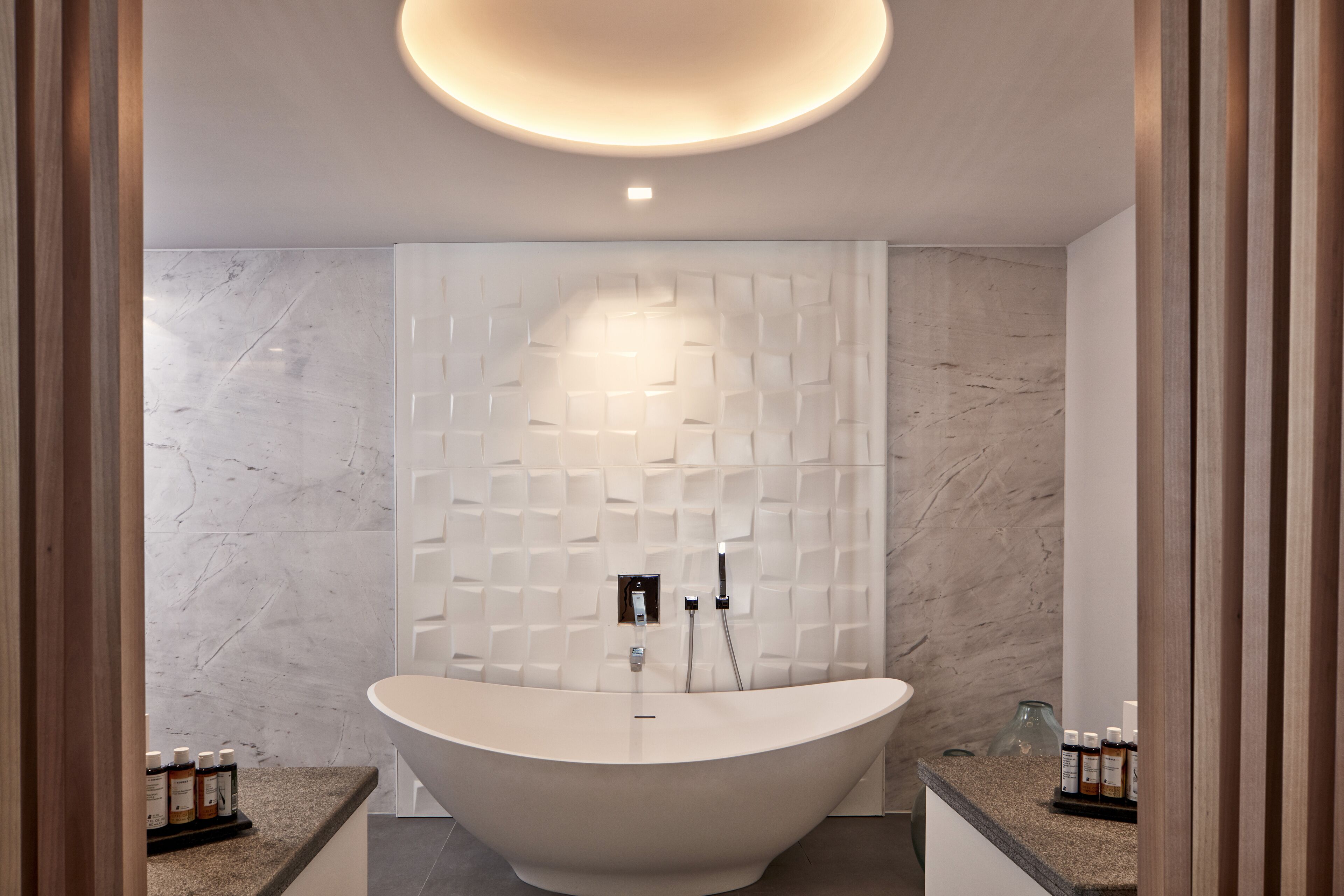 A modern bathroom with a freestanding bathtub, marble walls, and a recessed ceiling light.