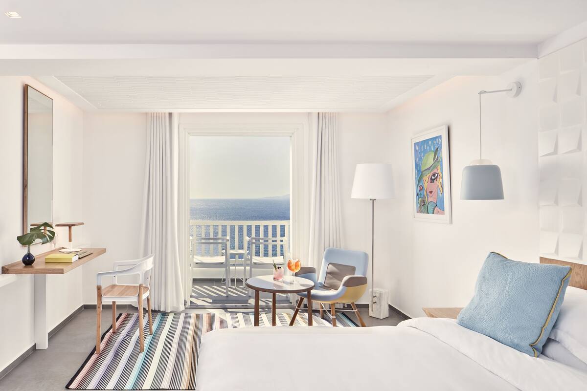 Ein Hotelzimmer mit Bett, Schreibtisch mit Stuhl, einem kleinen Tisch mit Getränken, einem Sessel und einem Balkon mit Meerblick. An der Wand hängt ein Bild.