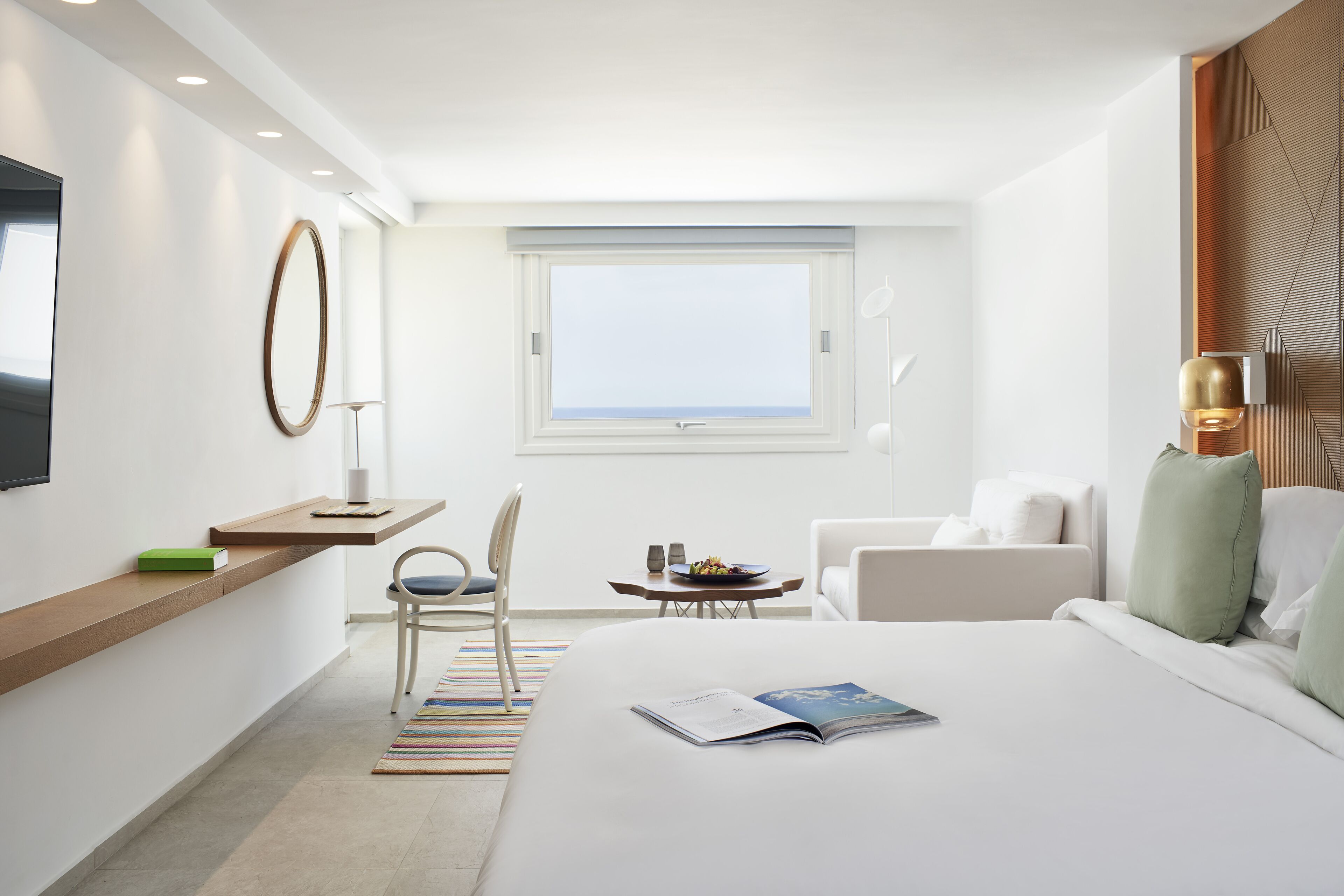Una habitación de hotel moderna con una cama grande, un escritorio con una silla, una mesita con un frutero, y vista al mar a través de una ventana.
