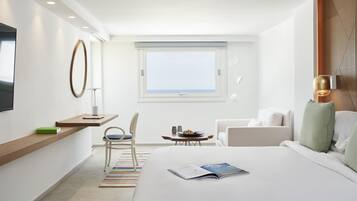 Una habitación de hotel moderna con una cama grande, un escritorio con una silla, una mesita con un frutero, y vista al mar a través de una ventana.