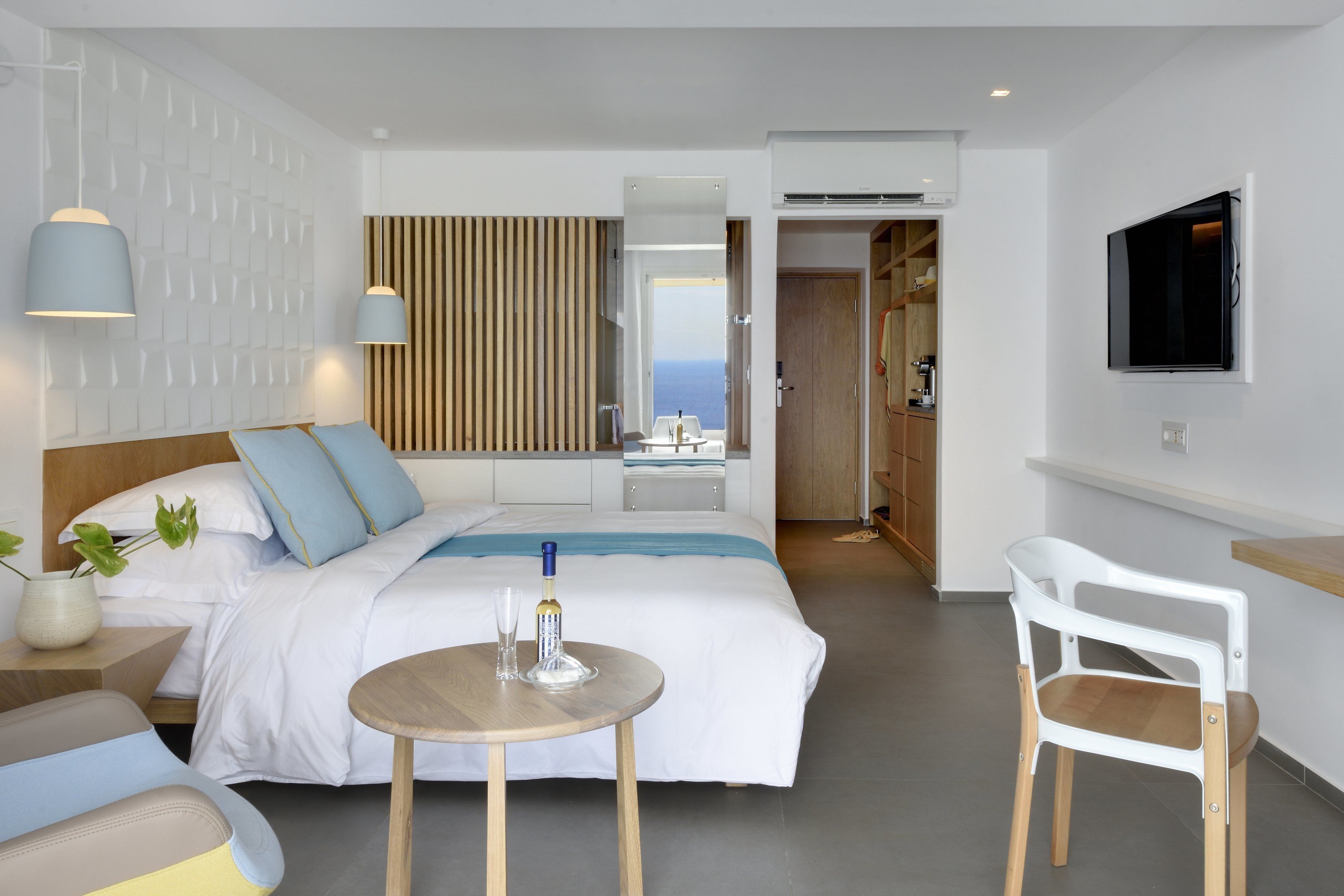 A modern hotel room with a large bed, bedside table, and a small round table with a bottle and glass. There is a TV mounted on the wall and a view of the sea through the window.