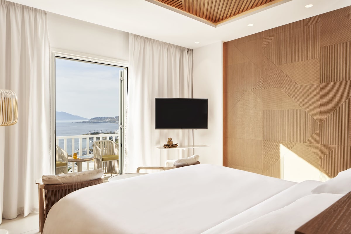Ein Hotelzimmer mit einem großen Bett, einem Fernseher, einem Balkon mit Meerblick und einer Wand mit Holzakzenten.