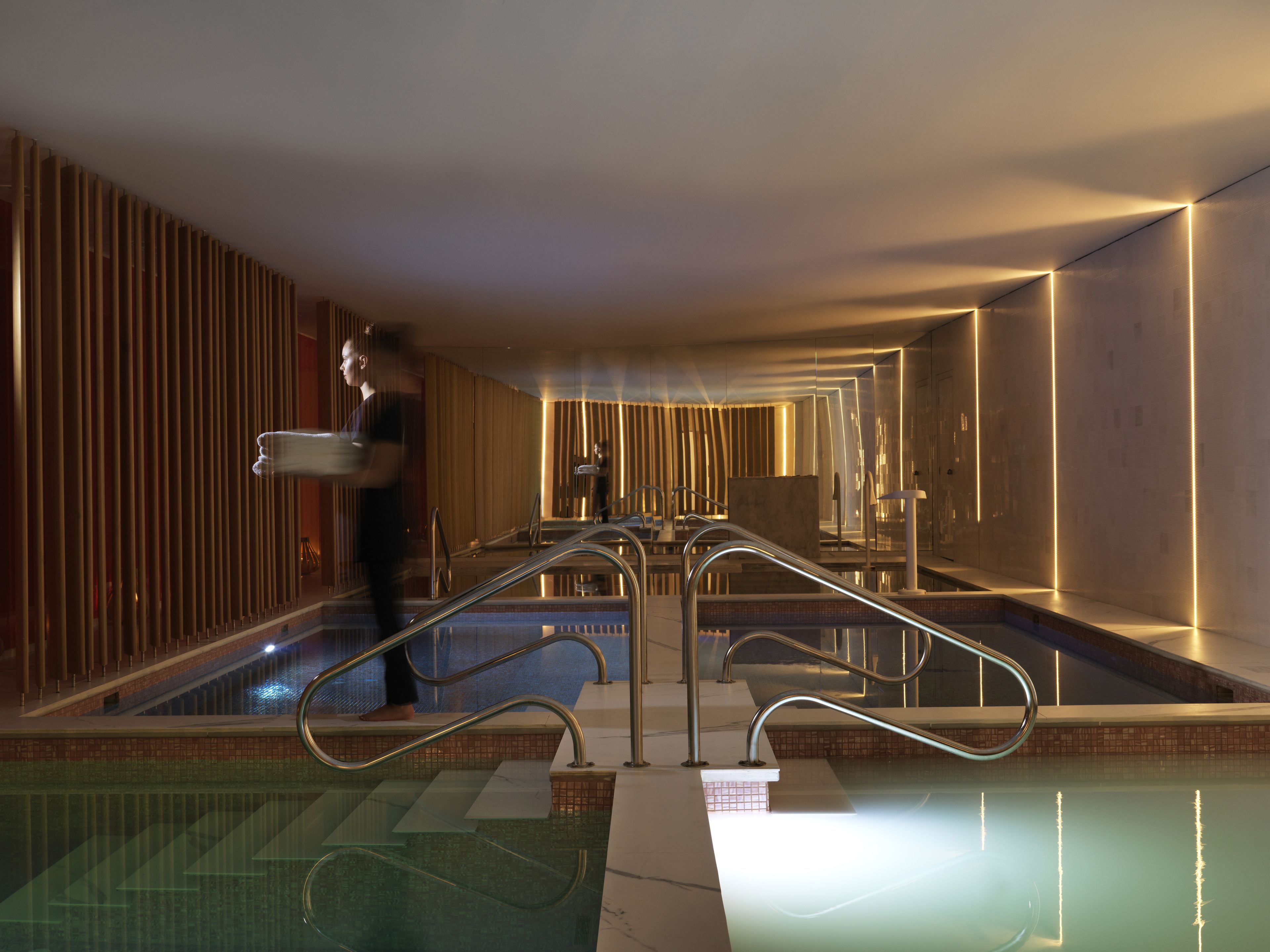 Une piscine intérieure moderne, dotée d’une paroi en verre, de rampes métalliques et d’une personne portant un plateau.
