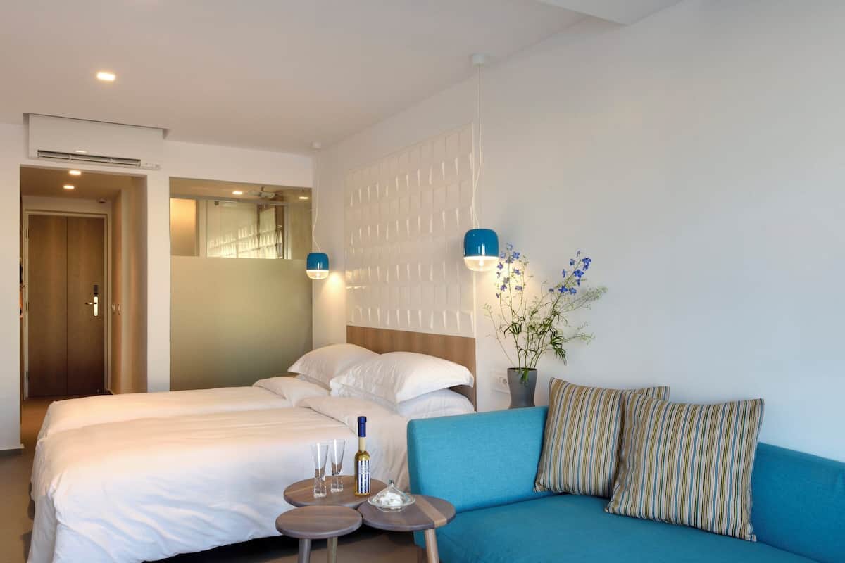 Ein Hotelzimmer mit Bett, Sofa, einem kleinen Tisch mit Gläsern und einer Flasche sowie einer Vase mit Blumen.